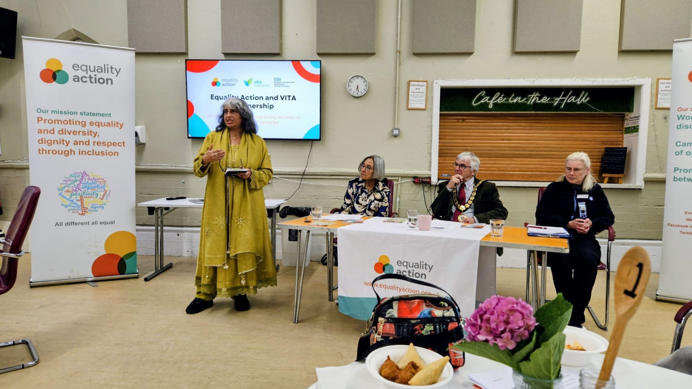 Equality Action trustee Abida Akram stands and speaks at the AGM wearing a vibrant yellow outfit.