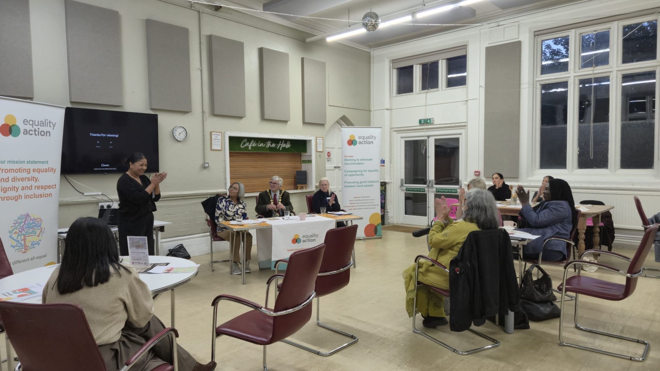 The audience at Equality Action’s AGM applauds at the end of the session. Yasmin Ali, the newly elected Chair of Trustees, is seated at the front, smiling.